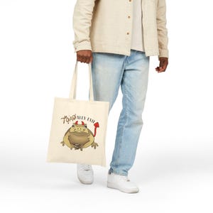 May include: A cream-colored tote bag featuring a cartoon illustration of a toad with devil horns and a tail. The text "TOADALLY EVIL" is above the toad. The bag is held by a person wearing light-wash jeans and a beige jacket.