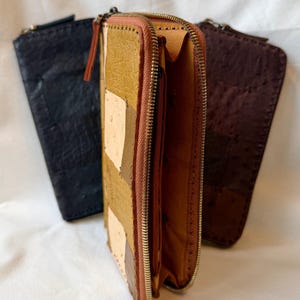 Op de afbeelding: Drie leren portemonnees in zwart, bruin en beige. De middelste portemonnee is open en toont de binnenkant. De voorste portemonnee heeft een patchwork ontwerp met een ritssluiting. De andere twee portemonnees zijn gesloten, met een textuur.