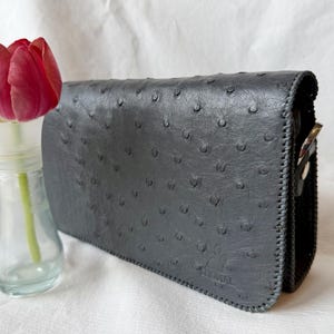 May include: A grey leather clutch bag with a textured surface and a stitched border. A red tulip in a clear glass vase is to the left of the bag. The bag has a silver clasp.