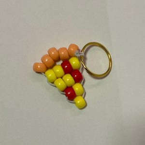 Op de afbeelding: Een sleutelhanger in de vorm van een pizzapunt gemaakt van kleurrijke kralen. De sleutelhanger heeft oranje, gele en rode kralen, die lijken op een pizzapunt. Een goudkleurige metalen ring is bevestigd voor eenvoudige bevestiging.