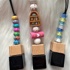 May include: Three car air fresheners with black square bottles, wooden tops, and beaded accents. One has colorful beads, another a bookshelf charm, and the third has clear and blue beads. Each hangs from a black cord.