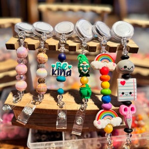 May include: A collection of retractable badge holders with various decorative charms. Charms include beaded strands, a gnome, a rainbow, and a notepad with a heart. The badge reels are displayed on a wooden stand.