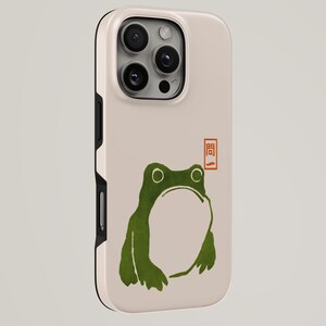May include: A beige smartphone case featuring a green frog illustration and a small red rectangular stamp with the Japanese characters 問一. The case has a black side frame and a cutout for the triple camera lens array on the back.