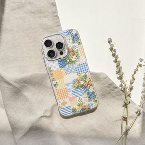 May include: A patterned phone case with a floral patchwork design. The case has a clear border and features blue, yellow, and white floral and gingham patterns. The phone case is on a beige linen cloth.