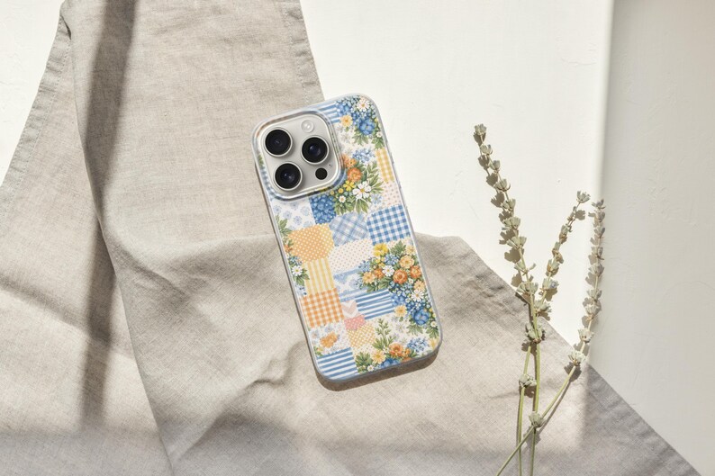 May include: A patterned phone case with a floral patchwork design. The case has a clear border and features blue, yellow, and white floral and gingham patterns. The phone case is on a beige linen cloth.