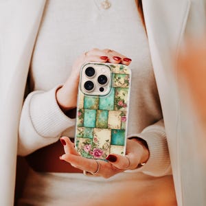 May include: A phone case with a patchwork design of teal and cream squares, adorned with floral motifs. The case has a clear border and a multi-lens camera aperture. The design evokes a vintage, feminine style.