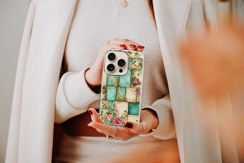 May include: A phone case with a patchwork design of teal and cream squares, adorned with floral motifs. The case has a clear border and a multi-lens camera aperture. The design evokes a vintage, feminine style.