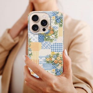 May include: A colourful patchwork phone case with floral and gingham patterns in blue, yellow, and white. The phone case is held in a person's hand. The case has a mix of floral patterns and gingham checks.