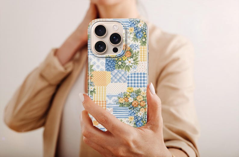May include: A colourful patchwork phone case with floral and gingham patterns in blue, yellow, and white. The phone case is held in a person's hand. The case has a mix of floral patterns and gingham checks.