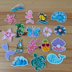 Puede incluir: Una colección de parches bordados con temas tropicales y acuáticos, que incluye una sirena, una tortuga marina, una piña, estrellas de mar, conchas, palmeras, una mariposa, una flor, una ola, una botella de Love Potion y una cola de ballena.