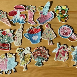 Op de afbeelding: Een verzameling geborduurde patches met grillige ontwerpen. Inclusief een ijsje, paddenstoelen, een fee, een laars, een theekopje met aardbeien, bloemen en een konijn. De patches zijn in verschillende kleuren.