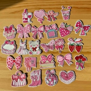 May include: A collection of pink embroidered patches featuring various designs. These include bows, hearts, a teddy bear, a dress, a cowboy boot, a purse, a strawberry, a rose, a wine bottle, and a drink.