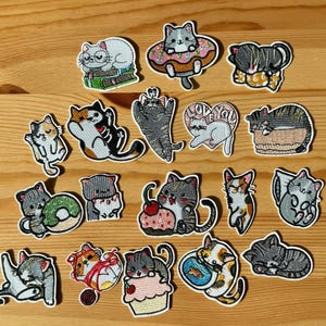 May include: A selection of embroidered cat patches in diverse poses and designs. Cats are shown with donuts, cupcakes, books, and hearts. The patches feature a variety of colours, including grey, orange, white, and black, with white borders.