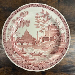 May include: A decorative ceramic plate with a scenic illustration in red and white. The design features a river scene with buildings, bridges, and boats. The plate has a scalloped edge and a detailed border.