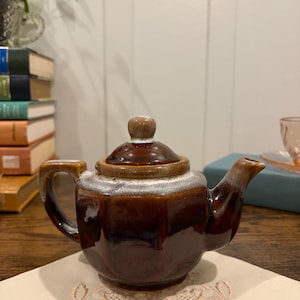 May include: A white ceramic mug with a black handle and the text "But First, Coffee" in a bold, black font. The mug is set against a blurred background, suggesting a cozy setting.