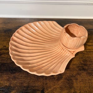 May include: A peach-colored ceramic serving dish shaped like a large scallop shell with a smaller, integrated bowl attached to one side. The surface features a glossy finish and prominent, radiating ridges that mimic the texture of a seashell.