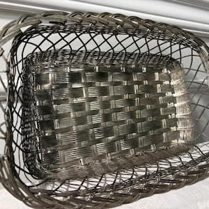 May include: A silver-toned metal basket featuring a rectangular base with a woven pattern and rounded, open-wire sides. The structure is crafted from thin, interconnected metal strips that form a lattice design, creating a deep, oval-shaped container.