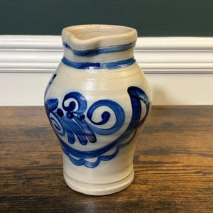 May include: A beige ceramic pitcher featuring a hand-painted blue floral pattern with swirling designs. The vessel has a wide, rounded body, a narrow neck, and a small pouring spout at the top rim, finished with a smooth, glazed surface.