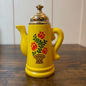 May include: A small, yellow ceramic teapot with a gold-colored lid and handle. The teapot is decorated with a floral design in red and green. The teapot is approximately 5 inches tall.