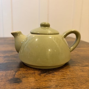 May include: A small, olive green ceramic teapot with a matching lid and curved handle. The teapot has a rounded body and a spout for pouring. It sits on a wooden surface.