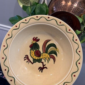 May include: A cream-colored ceramic bowl with a rooster illustration in the center. The rooster is depicted in yellow, green, and red. The rim of the bowl has a wavy green design with red dots. Kitchenware.