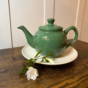May include: A green ceramic teapot with a matching lid and handle sits on a white saucer. A sprig of white flowers and green leaves rests on the dark wooden surface, adding a touch of nature to the scene.