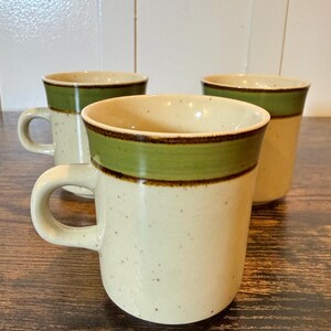 May include: Three ceramic mugs with a speckled beige body and a green band near the rim. The mugs have a brown accent line and a curved handle. The mugs are on a dark wood surface.