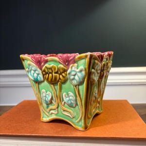 May include: A square ceramic planter featuring a light green base with raised, relief-style floral motifs in shades of pink, yellow, and light blue. The vessel has a flared rim and a scalloped base, with a glossy, crackled glaze finish throughout.