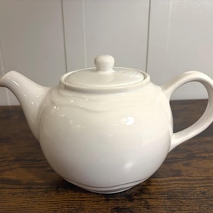 May include: Cream-colored ceramic teapot with a rounded body, curved spout, and a loop handle. The lid has a small knob for easy removal. The teapot is sitting on a dark wooden surface.