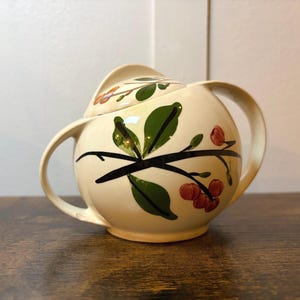 May include: Cream-colored ceramic sugar bowl with a lid and two handles. The bowl is decorated with a black branch design, green leaves, and red berries. The sugar bowl is sitting on a wooden surface.