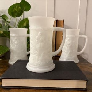 May include: Three white milk glass beer steins with handles, each featuring a detailed relief design. The steins are arranged on a dark book, with greenery and books in the background. The steins are approximately 6 inches tall.