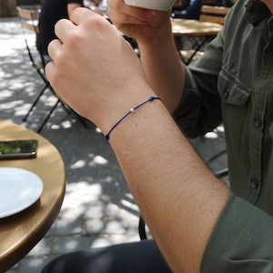 Könnte beinhalten: Ein dunkelblaues Armband mit einer kleinen silbernen Perle. Das Armband ist an beiden Enden mit Knoten versehen. Das Armband wird am Handgelenk getragen.