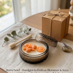 Op de afbeelding: Een sojawas kaars in een getextureerde glazen container met oranje bloem- en zonnebloemontwerpen. De kaars staat op een linnen oppervlak met een houten honingdipper en een kleine geschenkdoos vastgebonden met touw. Tekst op de afbeelding luidt: "Handgemaakt in Zweden - Premium Sojawas - Exotische bloemontwerpen - Milieuvriendelijk."