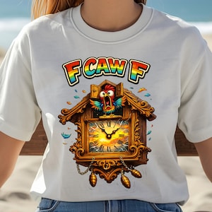 May include: White t-shirt featuring a colorful cartoon cuckoo clock design. The clock has a detailed wooden frame, a yellow clock face, and a red bird popping out. The text "F CAW F" is above the clock in rainbow colors.