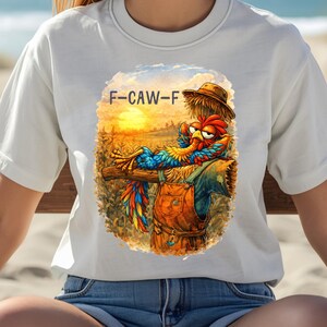 May include: White t-shirt featuring a colorful rooster illustration with the text "F-CAW-F". The rooster is depicted wearing overalls and a straw hat, set against a sunset backdrop.