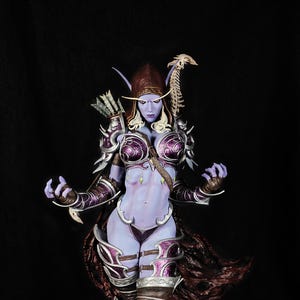 Op de afbeelding: Gedetailleerd beeldje van een fantasiekarakter met paarse huid, gekleed in een sierlijk harnas en een capuchon. Het figuur poseert met uitgestrekte armen, met een boog en pijlen. Het harnas is geaccentueerd met zilveren en paarse details, op een rotsachtige basis.