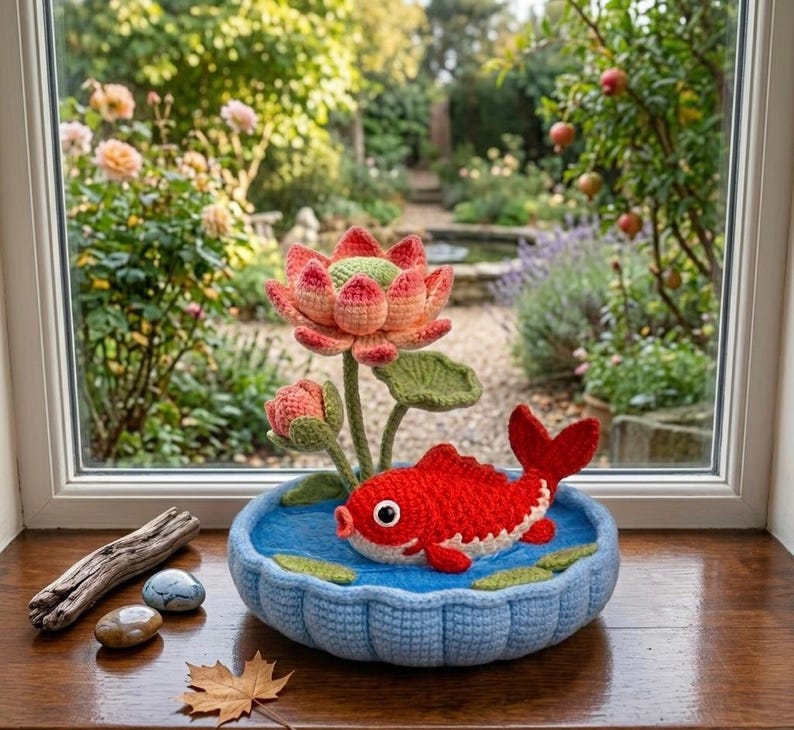 Puede incluir: Escena de ganchillo hecha a mano con un pez koi rojo y blanco en un estanque azul con una flor de loto. La escena est&aacute; ambientada en un cuenco azul tejido a ganchillo, con nen&uacute;fares verdes, sobre un alf&eacute;izar de madera.