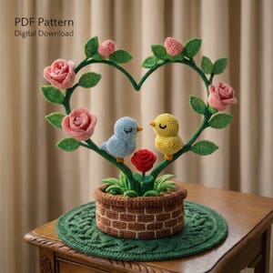 May include: A crocheted heart-shaped floral arrangement featuring pink roses, green leaves, and two small birds. The birds sit on branches above a brown basket-style pot. The text "PDF Pattern Digital Download" is in the upper left corner.