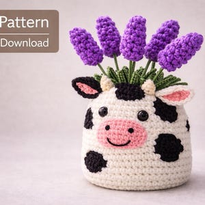 May include: A crocheted cow-shaped vase with black spots, a pink nose, and purple flower stems. The vase is filled with purple crocheted flowers. Text in the upper left corner reads "PDF Pattern Digital Download".