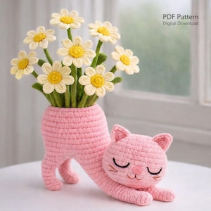 May include: A handmade pink crochet cat vase holding crochet daisies with yellow centers. The cat is in a stretching pose, with closed eyes and a smiling expression. The vase is a digital PDF pattern.