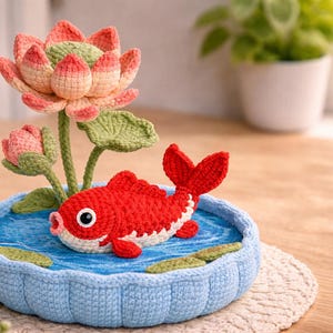 Puede incluir: Una pieza decorativa de ganchillo con un pez koi rojo en un estanque azul con una flor de loto. El pez tiene detalles blancos y ojos grandes. La flor de loto es rosa, verde y melocot&oacute;n. El estanque est&aacute; rodeado por un borde azul tejido.