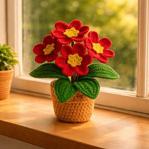 May include: A handcrafted crochet flower arrangement in a small pot. The flowers are red with yellow centers, green leaves, and a tan pot. The arrangement is placed on a wooden windowsill, with a window and greenery in the background.