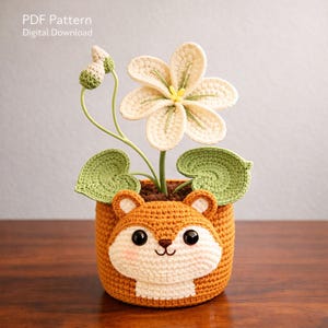Puede incluir: Una maceta de ganchillo con forma de ardilla, que contiene una flor de ganchillo con pétalos color crema y hojas verdes. La maceta es marrón con una cara blanca y ojos negros. El texto "PDF Pattern Digital Download" es visible.