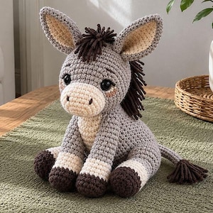 May include: A handmade crochet donkey plush toy in shades of gray, brown, and cream. The donkey has large, expressive eyes, long ears, and a fluffy mane and tail. The toy is sitting on a green surface.