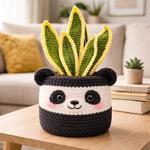 Puede incluir: Una maceta de panda de ganchillo hecha a mano con una planta de Sansevieria. La maceta es negra y blanca con un diseño de cara de panda, que incluye ojos negros, una nariz y mejillas rosadas. La planta tiene hojas verdes y amarillas.