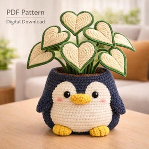Puede incluir: Una maceta de pingüino de ganchillo con hojas en forma de corazón. El pingüino es azul marino, blanco y amarillo, con mejillas rosadas. Las hojas son de color crema con ribete verde. El texto "PDF Pattern Digital Download" está en la esquina superior izquierda.