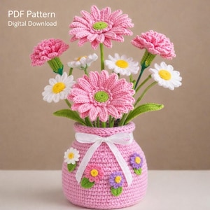 May include: A crocheted bouquet of pink gerbera daisies, carnations, and white daisies with yellow centres, arranged in a pink vase. The vase is adorned with crocheted flowers and a white ribbon. The text "PDF Pattern Digital Download" is in the upper left corner.