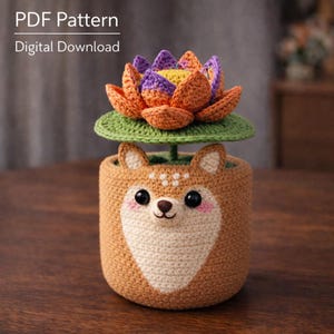 May include: Crocheted deer-themed planter with a colorful lotus flower. The planter is brown with a cream-colored face and pink cheeks. The lotus flower has orange, purple, and yellow petals. The text "PDF Pattern Digital Download" is at the top.