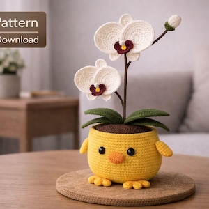 Op de afbeelding: Een gehaakte orchideeplant met witte bloemen en een gele pot in de vorm van een kuiken. De pot heeft kleine vleugels en voetjes. De tekst "PDF Pattern Digital Download" staat in de linkerbovenhoek.