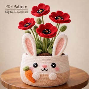 Könnte beinhalten: Ein gehäkelter Hasen-Blumentopf mit roten Mohnblumen. Der Hasen-Blumentopf ist weiß und beige mit einer Karotte, rosa Wangen und schwarzen Augen. Die Blumen haben schwarz-gelbe Zentren. Der Text "PDF Pattern Digital Download" befindet sich oben links.